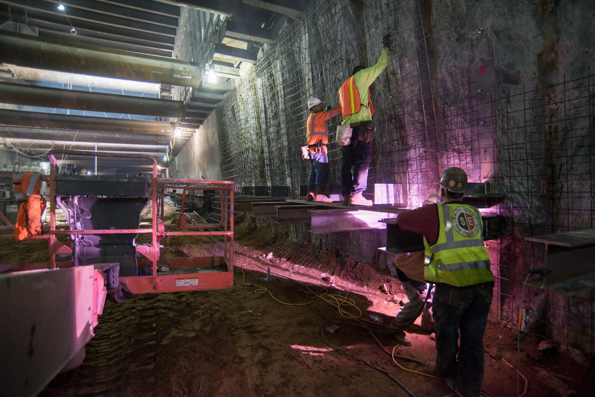 Workers in safety vests install steel mesh and brackets on station exterior walls at night.