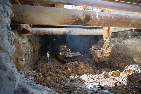 Construction excavation under bridge with heavy machinery and debris at street level.