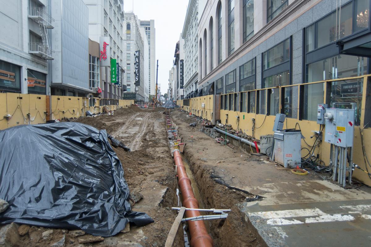 Construction trench with large sewer pipe along urban street surrounded by yellow barriers and buildings.