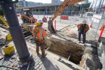 Workers in safety gear operate equipment at an open trench excavation on an urban street.