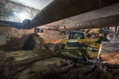 Bobcat loader removing excavated material beneath steel support beams in underground construction trench.