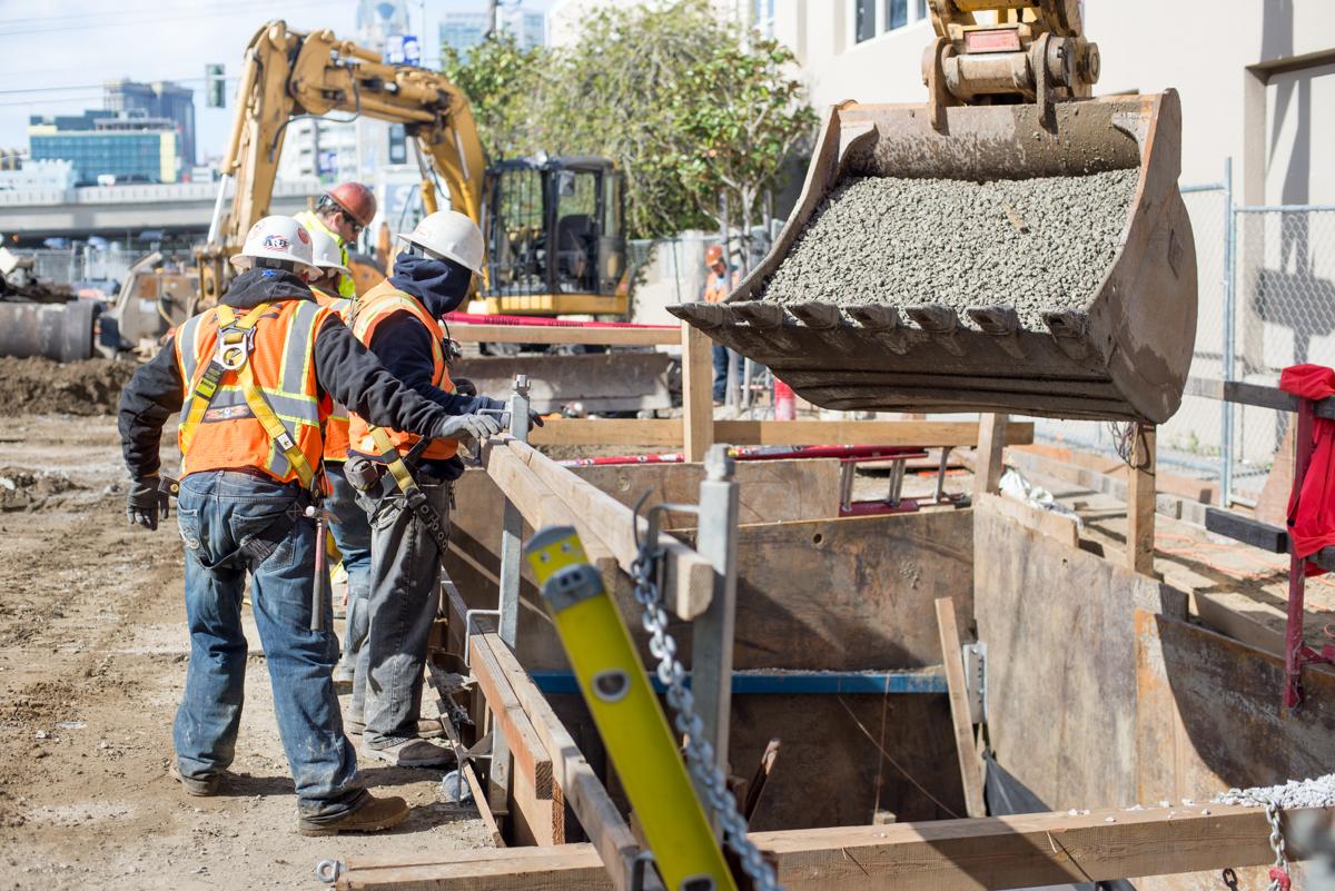Construction workers guide excavator bucket of gravel into trench during utility upgrade work on 4th Street.
