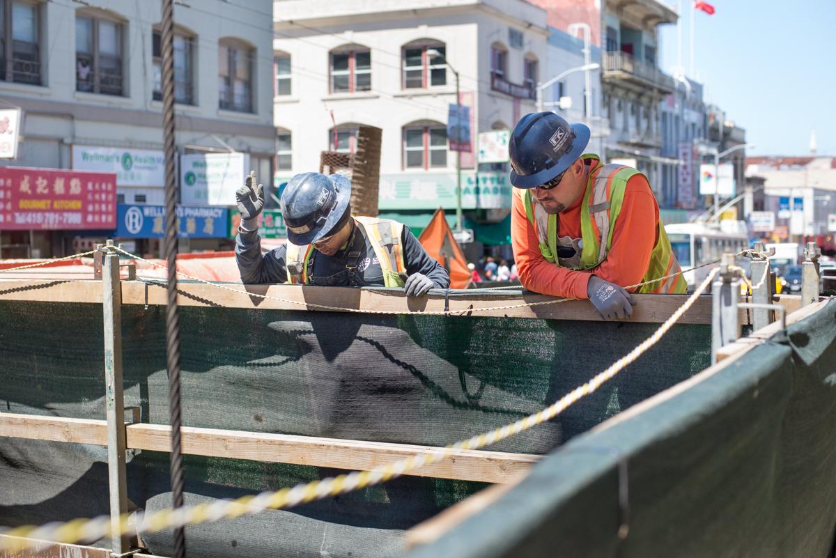 Three construction workers in hard hats and safety vests work at an underground excavation site in an urban area.