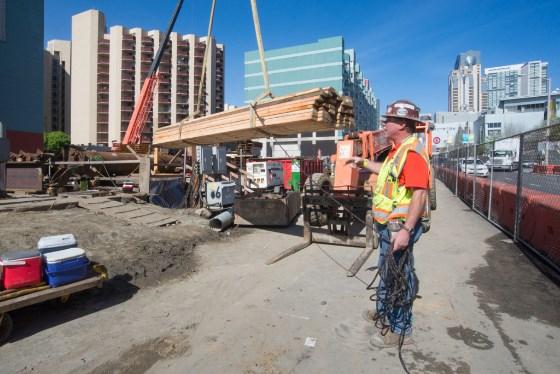 Construction worker in safety gear operating crane to lift wooden beam at urban transit station construction site.