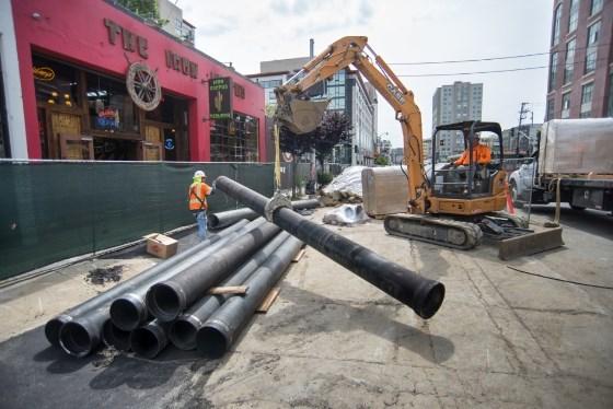 Construction crew with excavator positioning large pipes on 4th Street for Central Subway project installation.