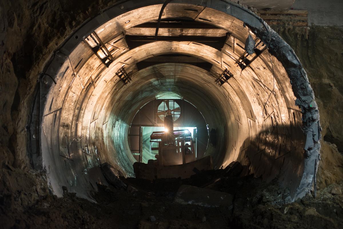 Interior view of Central Subway tunnel under construction, showing circular concrete tunnel with wooden bracing and equipment visible in distance.
