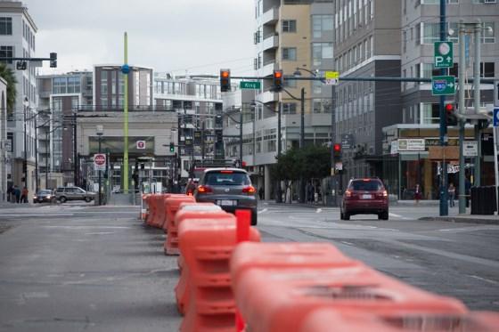 Red construction barriers line a San Francisco street intersection with traffic lights and downtown buildings.