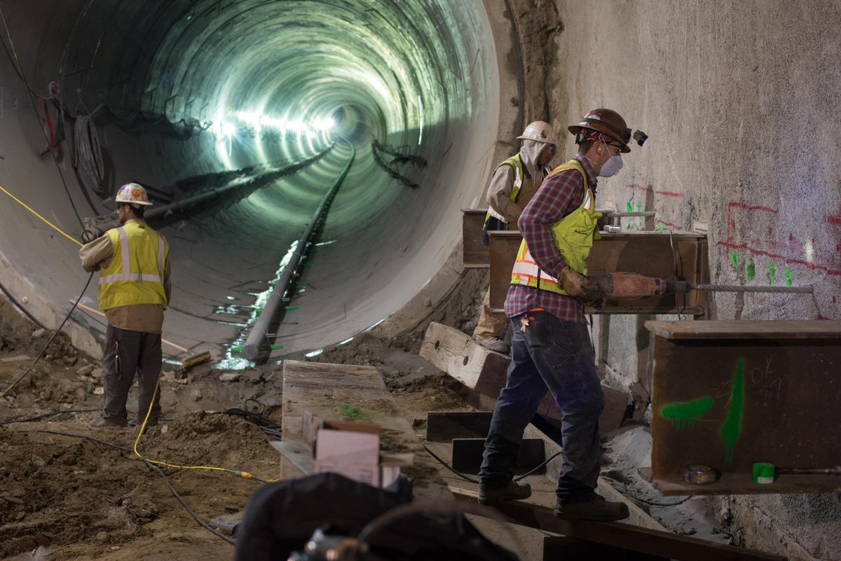 Workers in hard hats and safety vests inside a large tunnel during underground station construction and excavation.