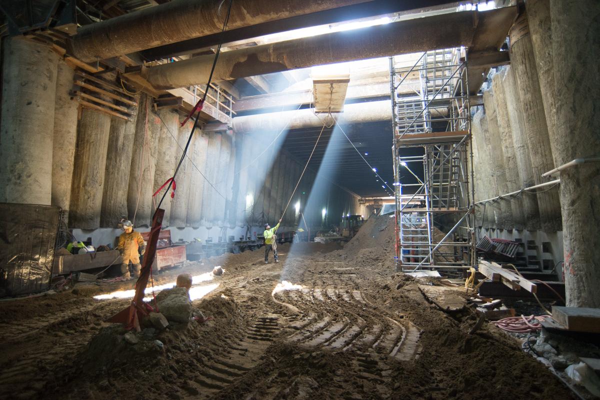 Construction workers excavate underground transit station between concrete support columns with scaffolding and equipment.
