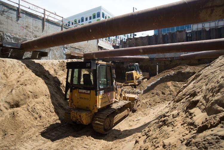 Caterpillar bulldozer excavating soil beneath steel bridge supports at transit station construction site.