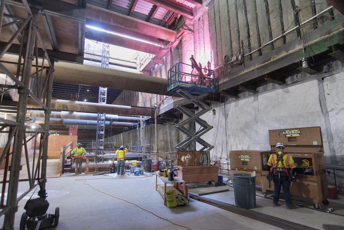 Construction workers in safety gear work inside a multi-level underground station with exposed structural elements and pink lighting.