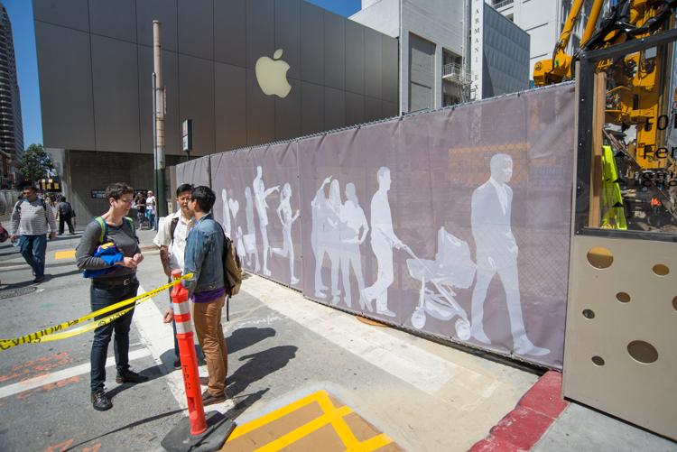 Construction barrier with white silhouette figures on brown mesh fencing along Stockton Street near Apple Store.