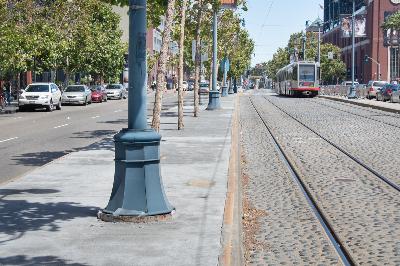 Streetcar tracks and sidewalk with utility pole on urban street lined with trees and parked cars.