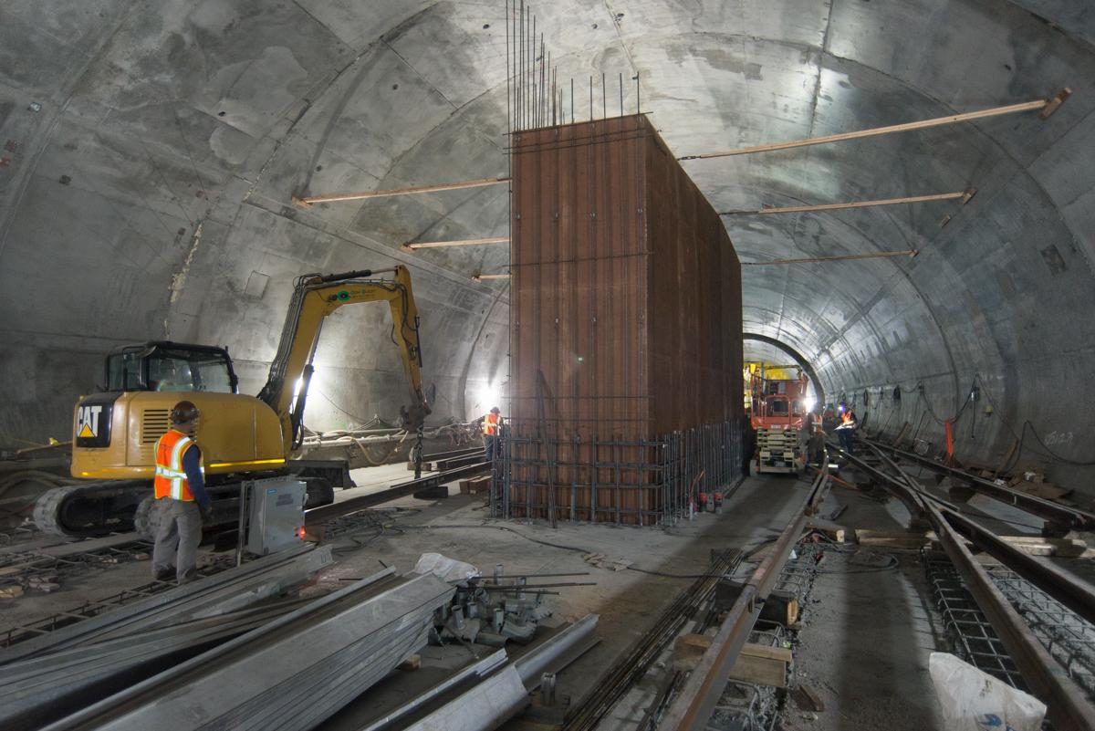 Construction workers and equipment install formwork for a large structure inside a subway tunnel with exposed concrete walls and railway tracks.