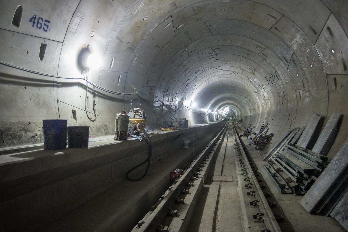 Underground transit tunnel under construction with railway tracks, concrete platform, and work equipment visible.