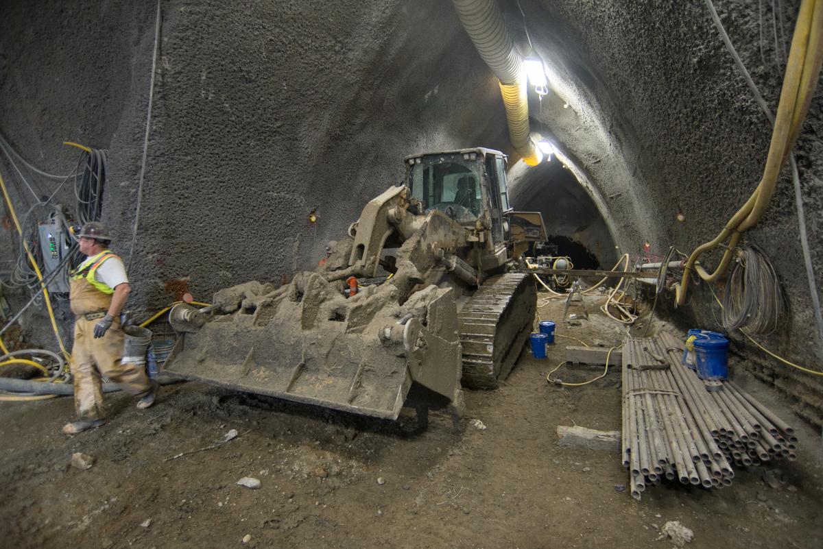 Construction equipment and workers inside underground tunnel during platform cavern excavation and shotcrete application.