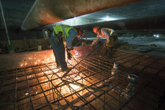 Workers in safety vests cutting rebar on a concrete construction site at night under a bridge.