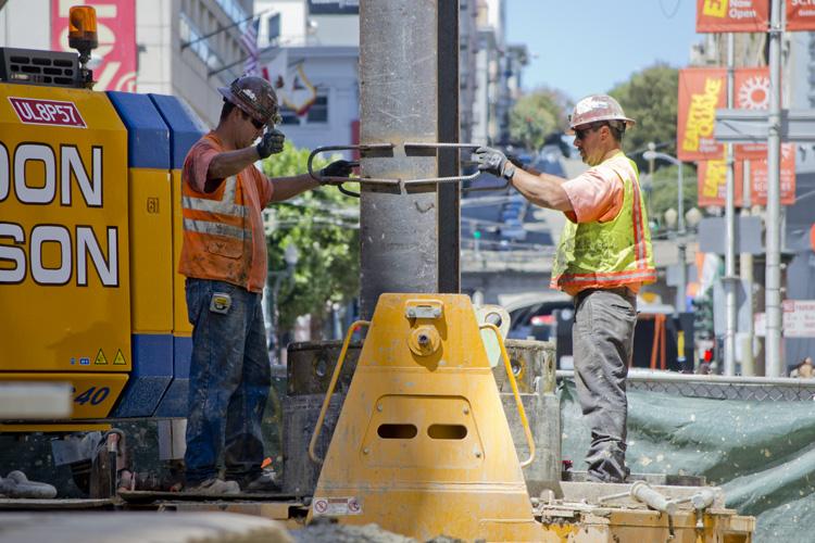 Two construction workers guide a steel beam being lowered into an underground excavation on a city street.