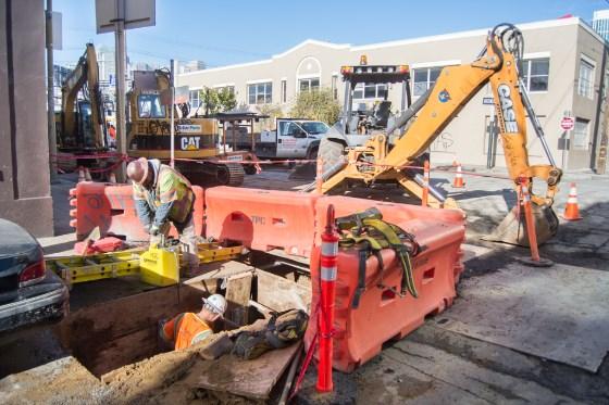 Construction crew working in street trench with excavator and safety barriers at transit project site.