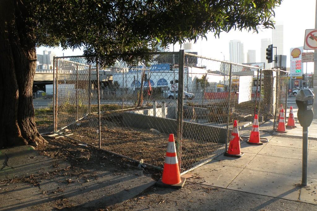Fenced construction site at urban street corner with traffic cones and city skyline in background.