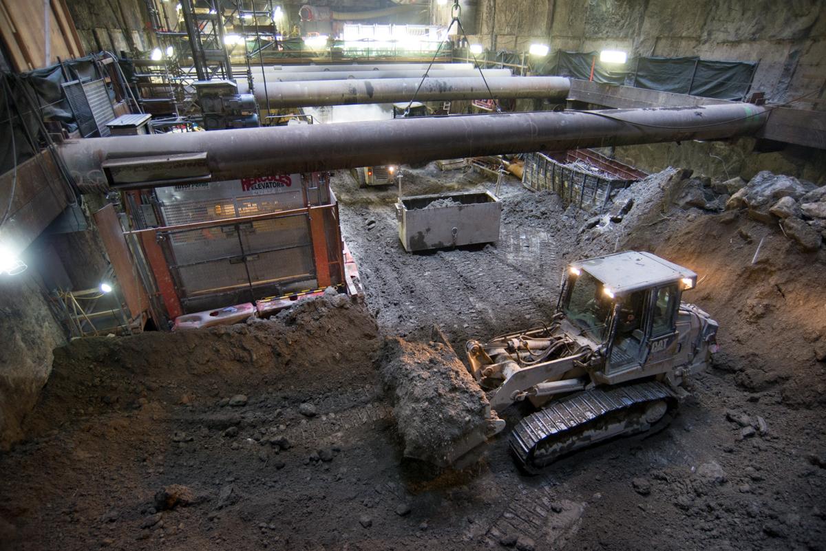 Underground construction site with excavator moving soil beneath large pipes and concrete structure.