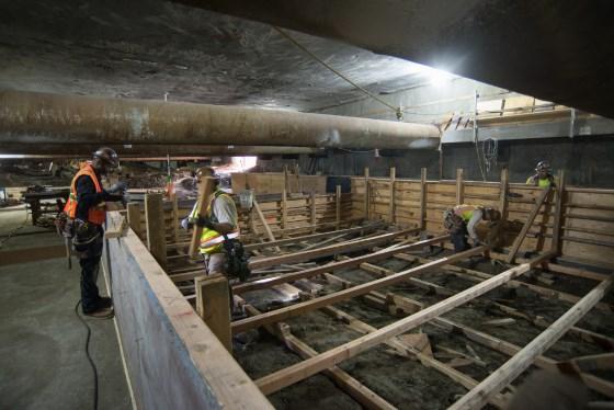 Workers in safety gear construct wooden formwork on a mezzanine level for concrete pouring at a transit station.