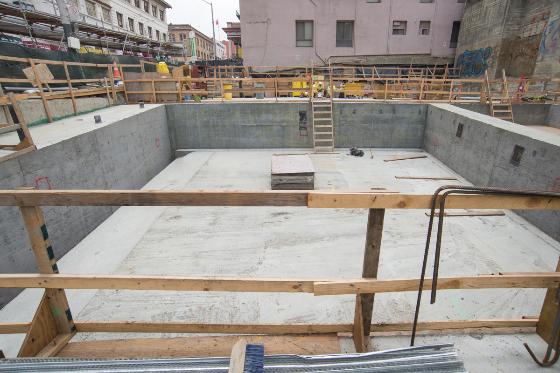 Underground station construction pit with concrete walls, wooden support beams, and metal railings, surrounded by urban buildings.