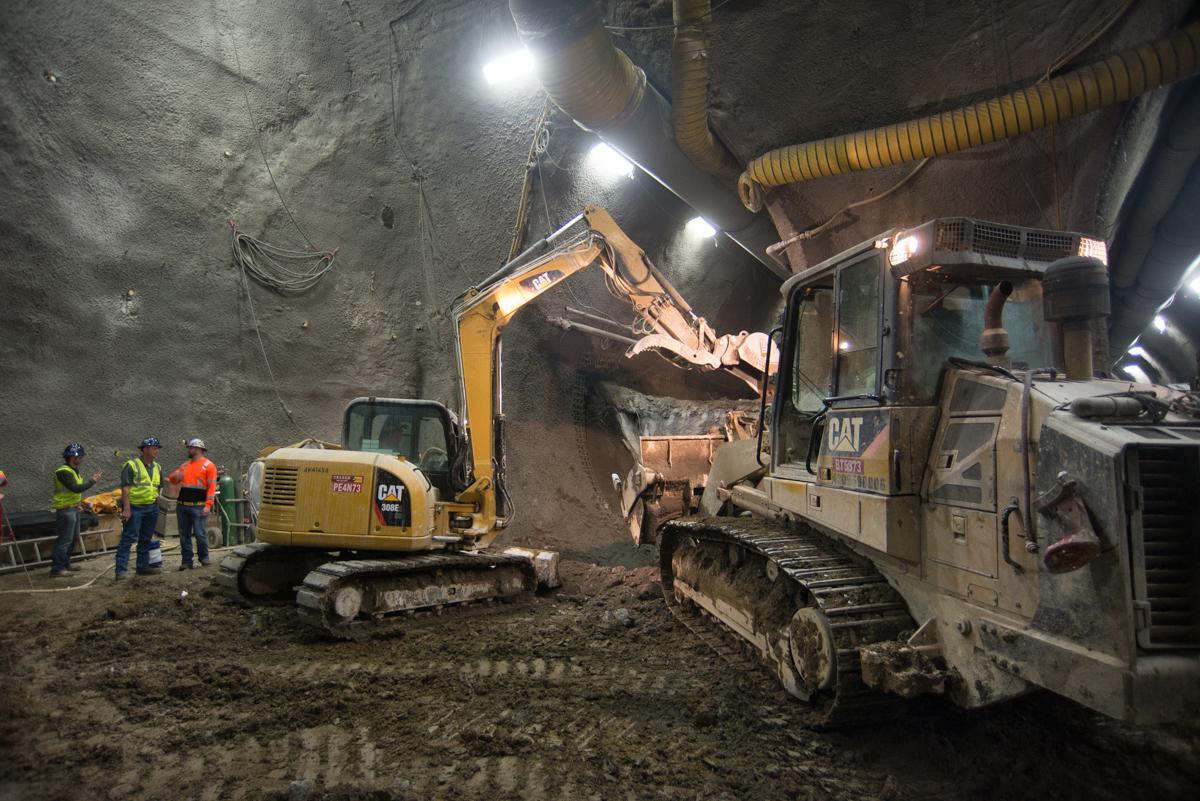 Excavator and bulldozer removing material from underground tunnel excavation at night with work crew and lighting.
