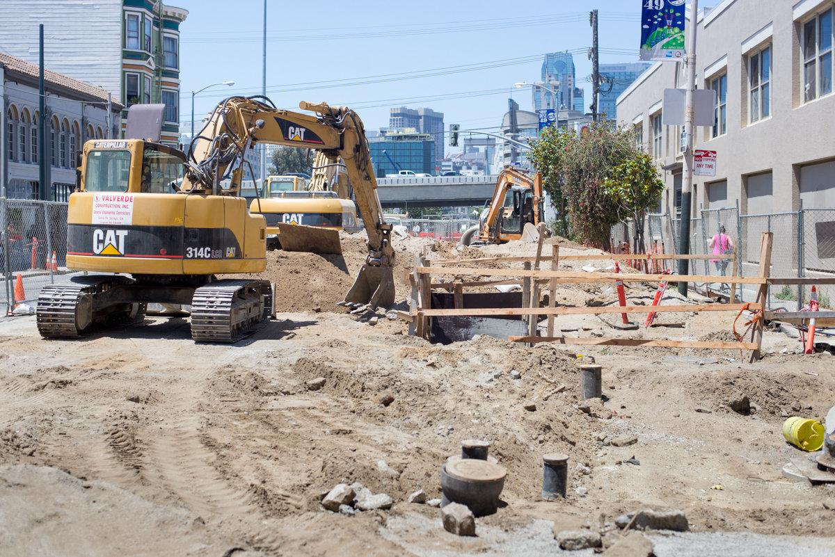 Construction excavators and equipment at Central Subway project site on 4th Street with city skyline in background.