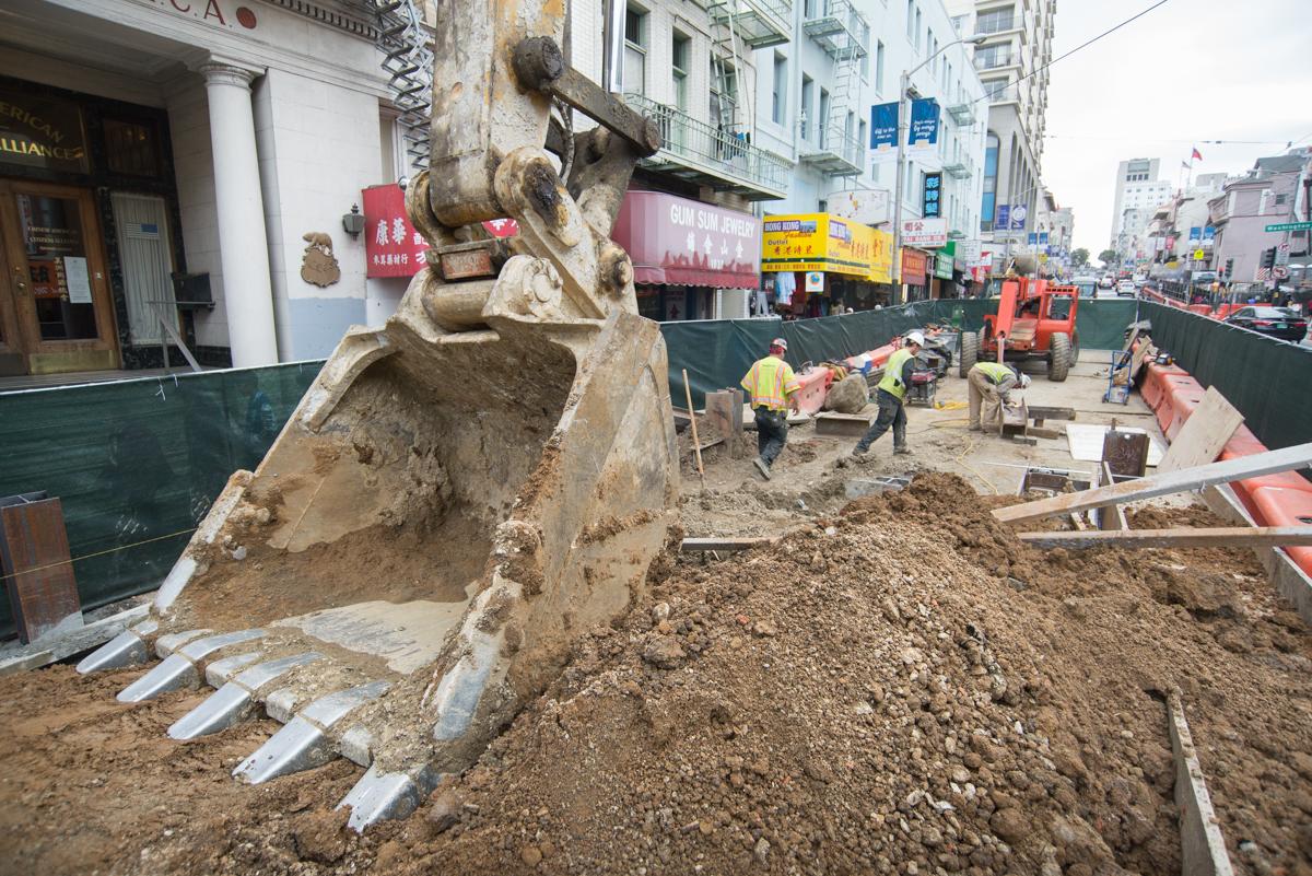 Excavator bucket positioned at construction site on Stockton Street with workers and equipment in background.