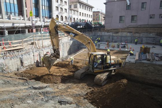 Excavator clears rocks and debris from construction site surrounded by urban buildings and safety barriers.