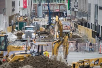 Construction site with yellow excavators and heavy equipment on urban street surrounded by buildings.
