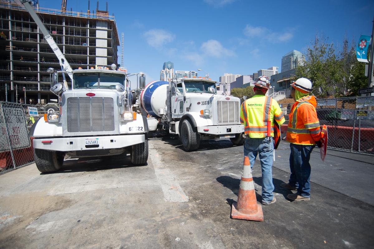 Construction workers in safety vests direct concrete mixer trucks at an urban transit construction site.