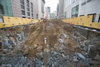 Construction excavation site with yellow barriers, exposed soil and concrete debris between urban buildings.