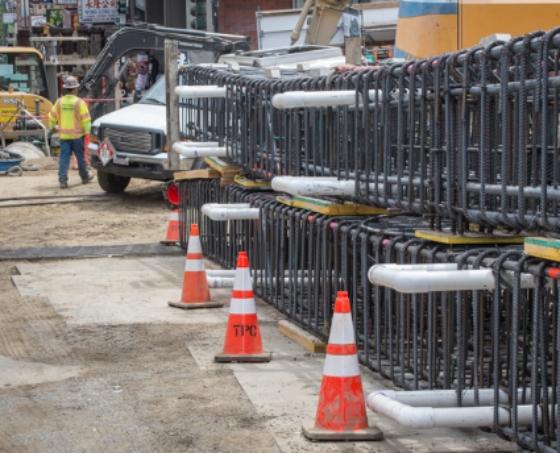 Rebar cages stacked at construction site with traffic cones and work vehicle nearby.