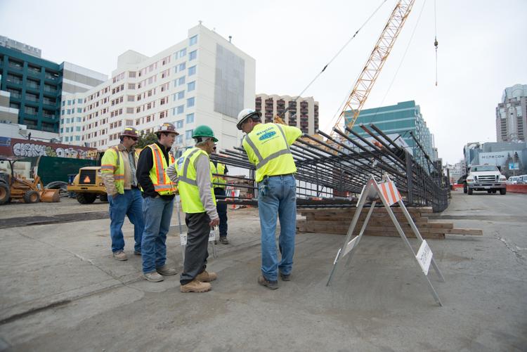 Construction workers in safety vests examine steel rebar cage at Yerba Buena/Moscone Station site with crane and urban buildings in background.