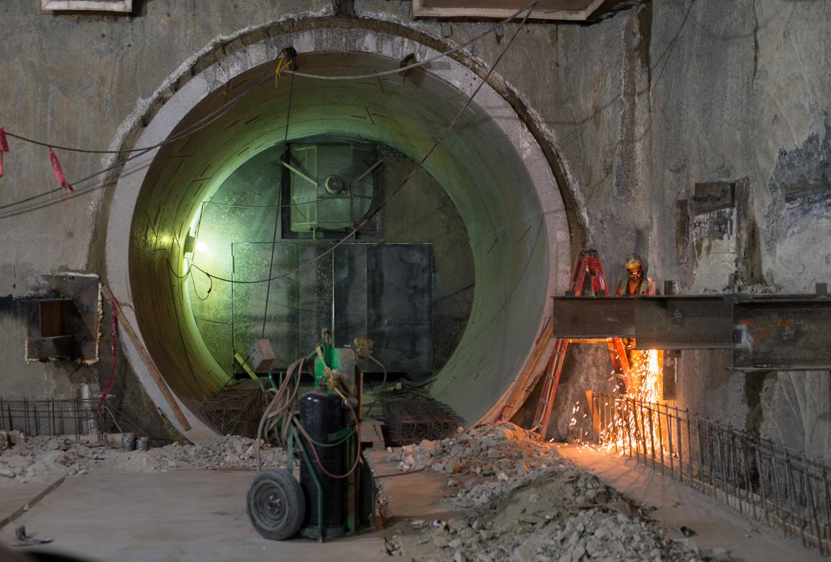Workers perform welding and construction inside a large circular tunnel section during transit station construction.