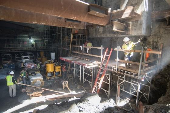 Construction workers install rebar at Chinatown Station headhouse with scaffolding and equipment visible.