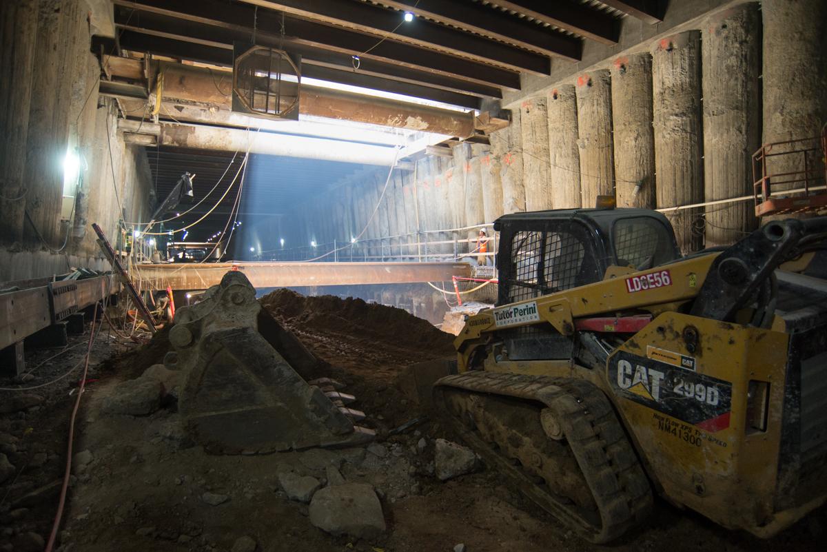 Yellow skid steer loader positioned in underground station box with excavated soil and temporary steel bracing visible.