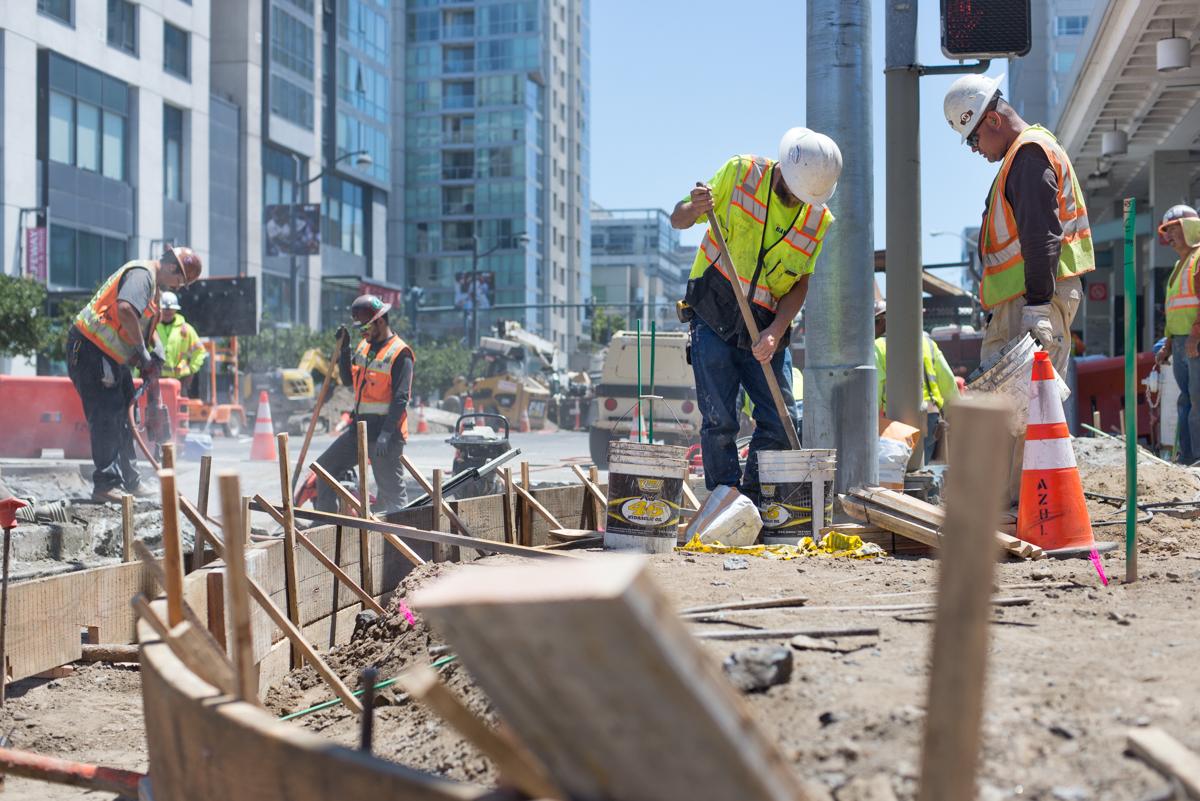 Construction workers jackhammer concrete around a manhole on an urban street surrounded by high-rise buildings.