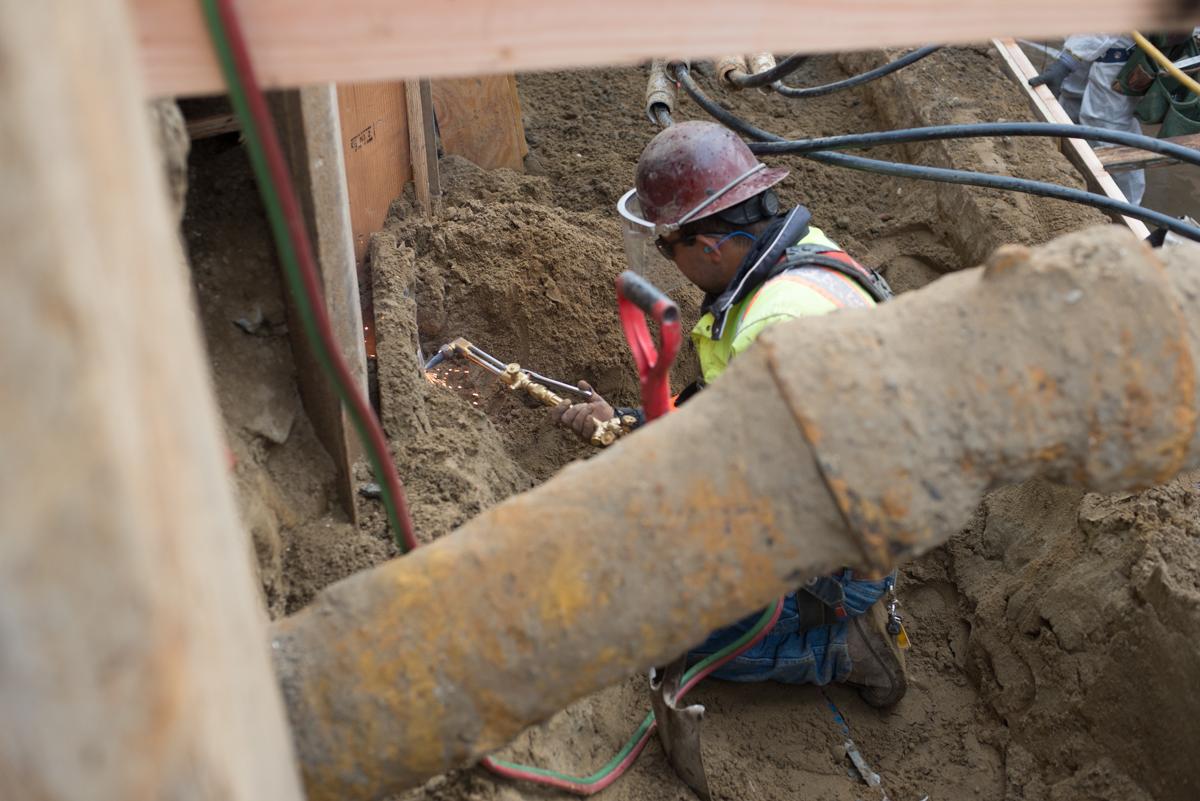 Worker in hard hat and safety vest performing utility work in excavated trench on 4th Street.