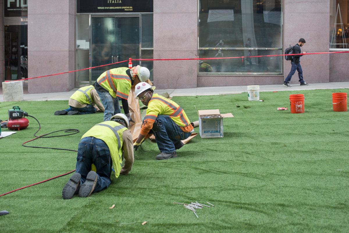 Workers in safety vests install artificial turf on an urban plaza in front of a building.