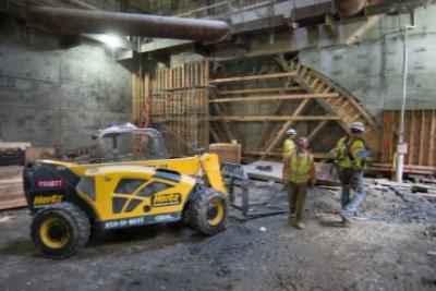Workers and equipment at underground transit station construction site with wooden framework and steel beams.