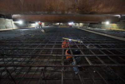 Worker assembling rebar cage under steel supports in underground trench excavation.