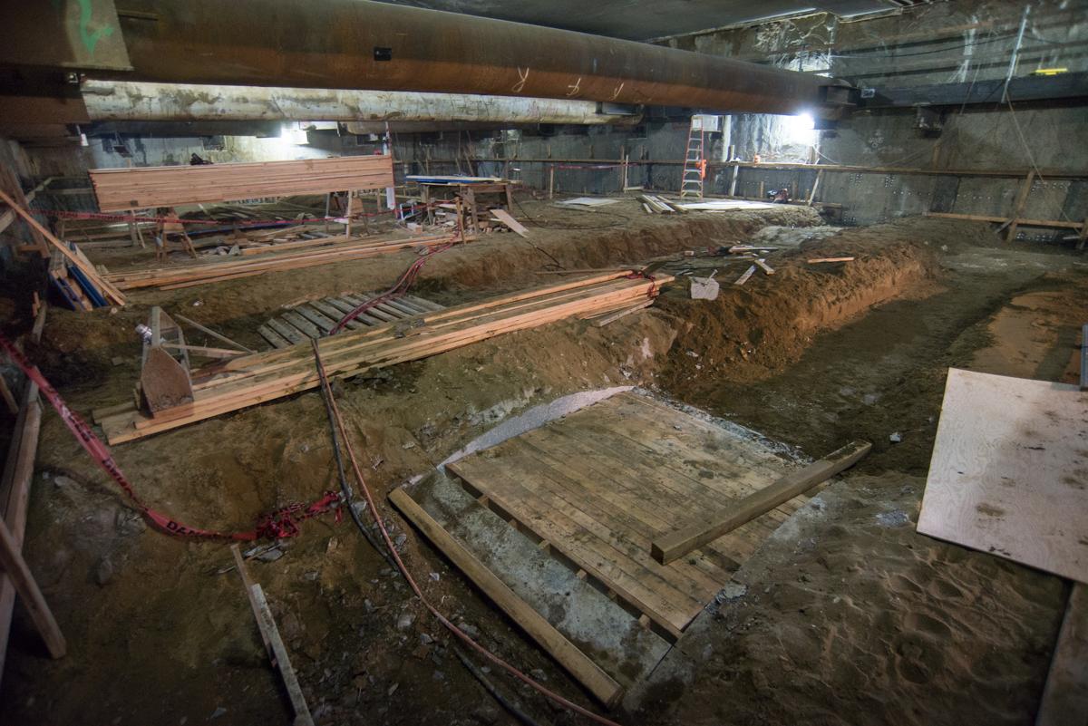 Construction site showing excavated trench with wooden formwork and scaffolding under concrete ceiling structure.