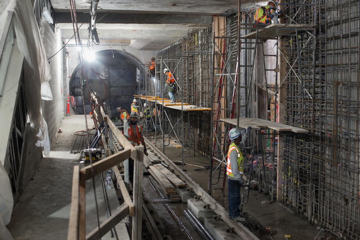 Workers in safety gear on a subway tunnel construction site with scaffolding and rebar framework visible.