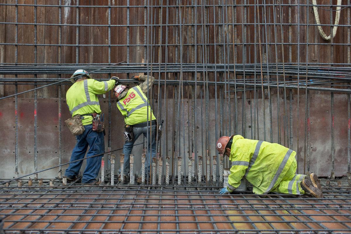 Three construction workers in yellow safety vests assemble steel rebar and scaffolding inside a station headhouse.