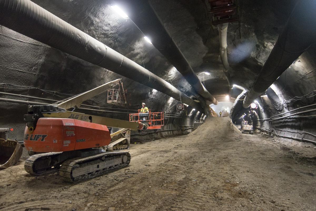 Underground tunnel construction with orange excavator and large pipes overhead under bright work lights.
