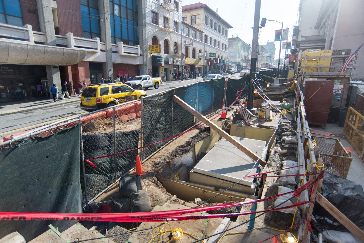 Construction site on Stockton Street showing excavation pit with rebar installation and equipment, yellow taxi passing above.