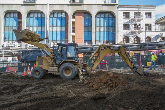 Backhoe excavating soil at Chinatown Station construction site with historic building facade in background.